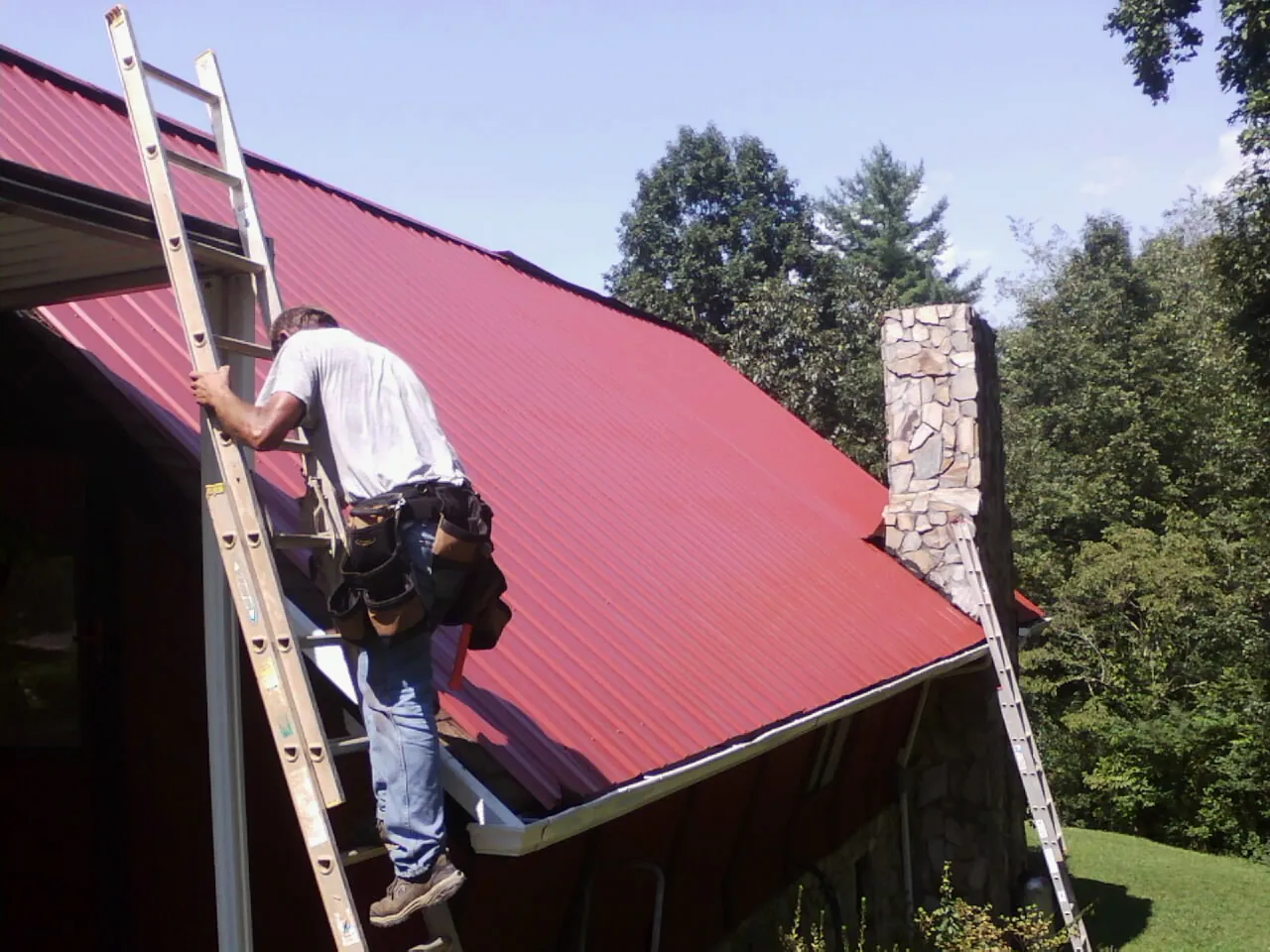 Roofer on a ladder beside a red metal roof and stone chimney — Soffit & Fascia Repair by Roofing Brothers in Big Sky